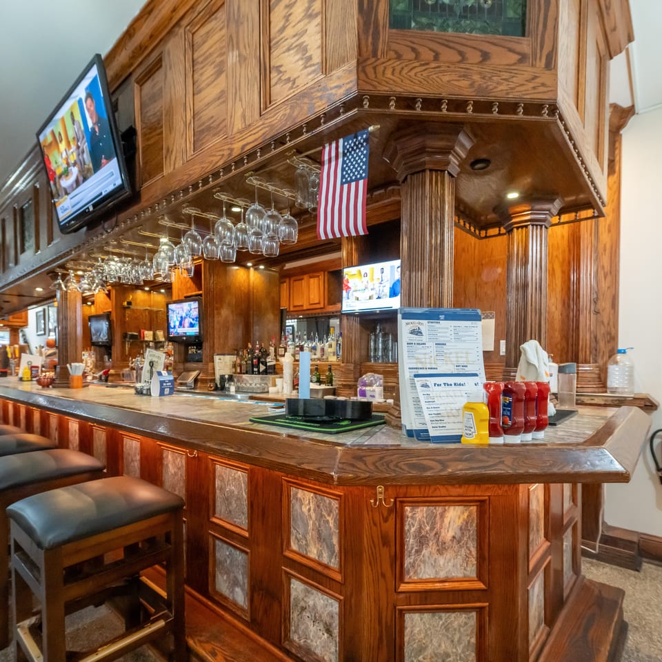 A richly wood-paneled restaurant bar features leather stools, hanging wine glasses, liquor bottles, televisions, and an American flag.