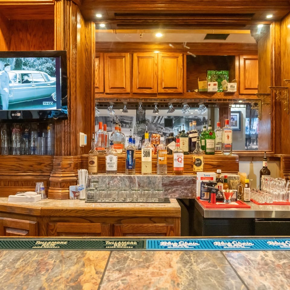 Wood-paneled restaurant bar stocked with liquor bottles and glassware, featuring a television and marble countertop.