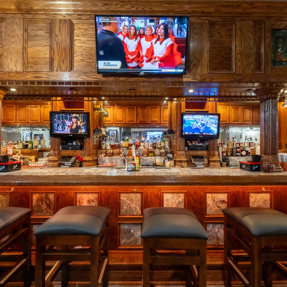 Elegant wood-paneled bar with liquor shelves, multiple televisions, hanging glassware, and cushioned stools.