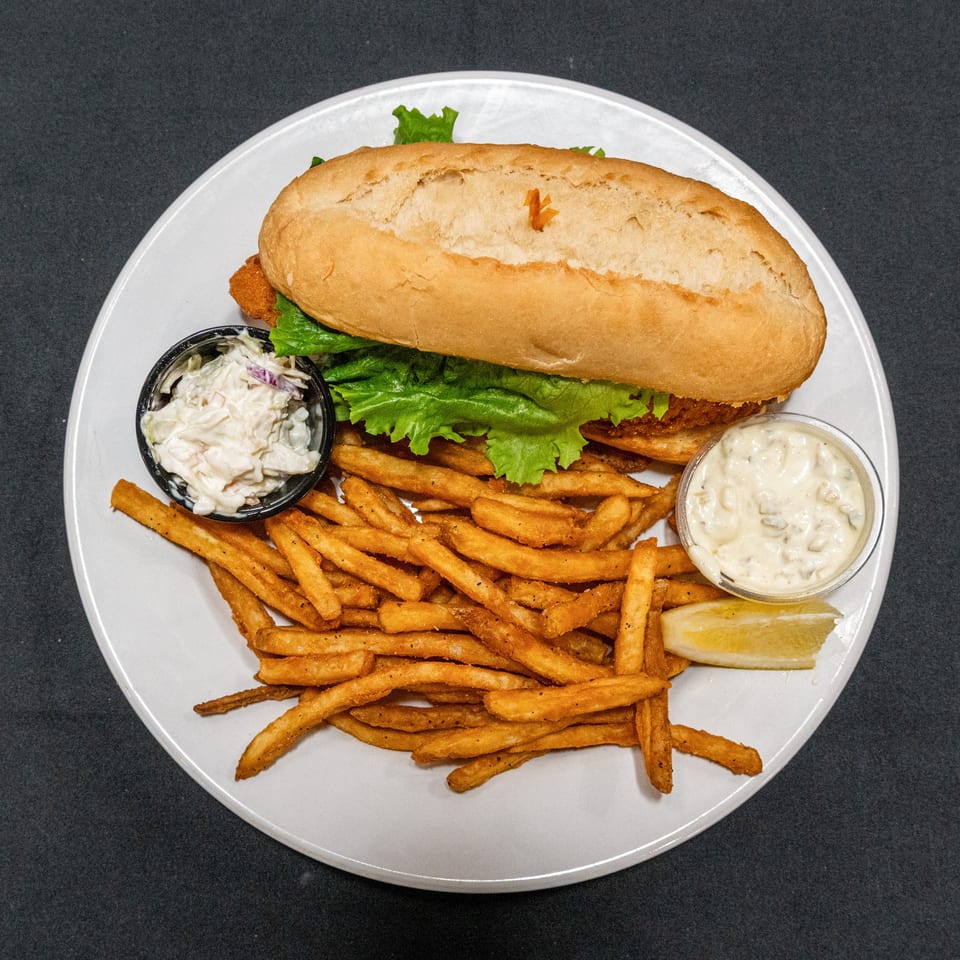 Crispy fried fish sandwich with lettuce, seasoned fries, coleslaw, tartar sauce, and lemon wedge on a white plate.