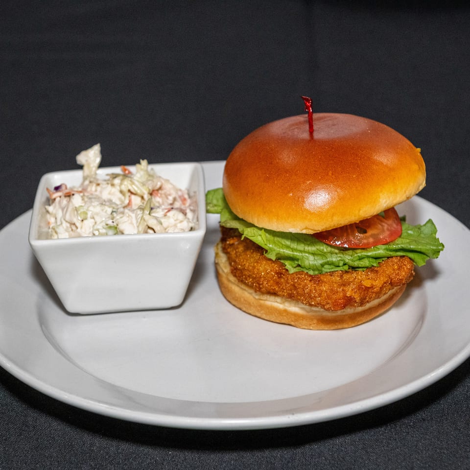 Crispy chicken sandwich with lettuce, tomato, and glossy brioche bun served beside creamy coleslaw on a white plate.