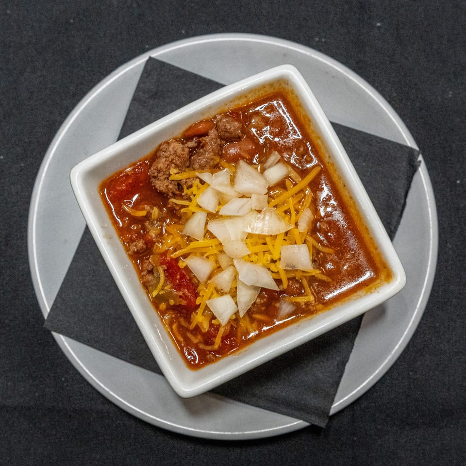 Overhead view of a bowl of chili topped with shredded cheddar cheese and diced onions on a plate with black napkins.