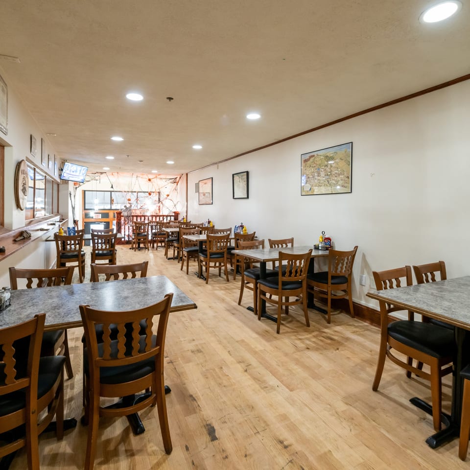 Bright, empty restaurant dining room with wooden tables and chairs, condiment baskets, framed artwork, and large windows.