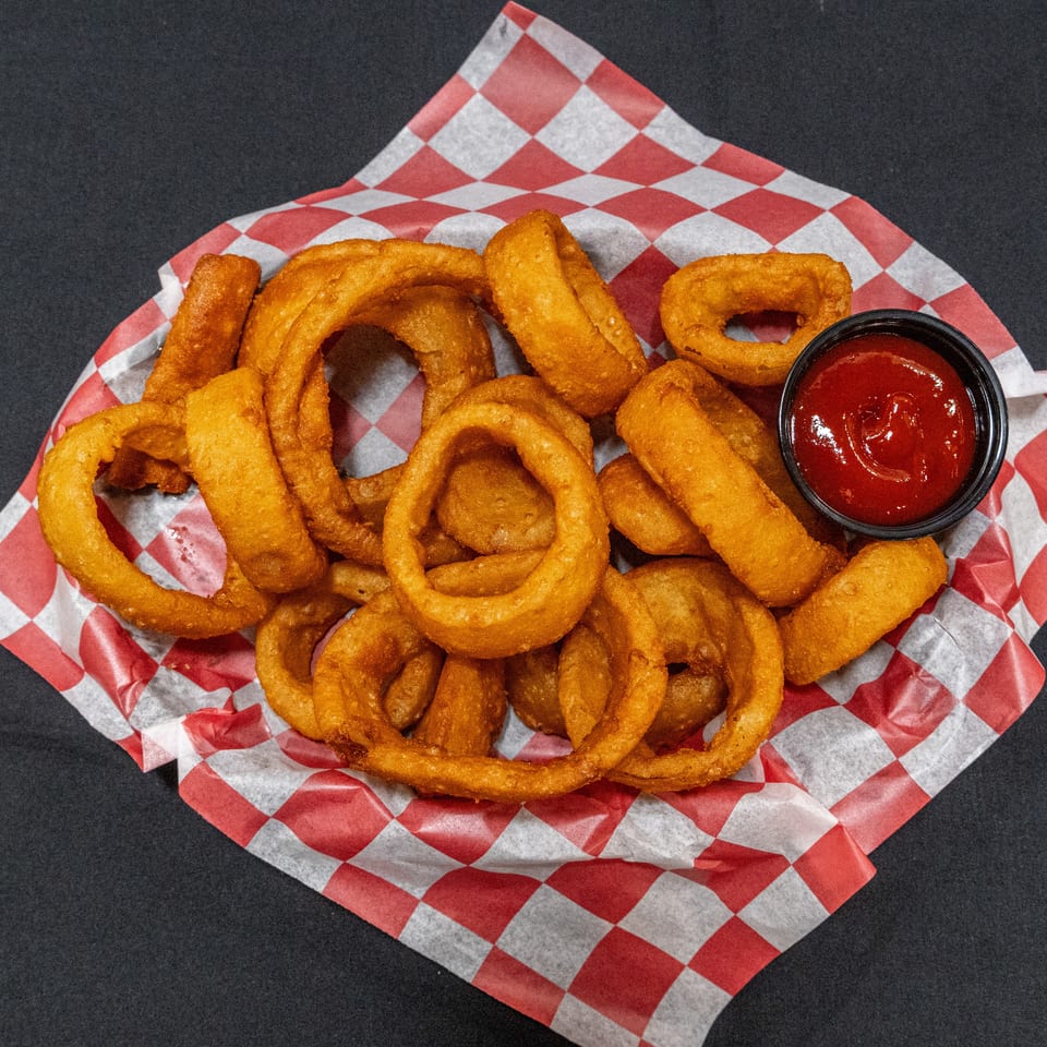 Golden fried onion rings served with ketchup in a black cup on red-and-white checkered basket liner.