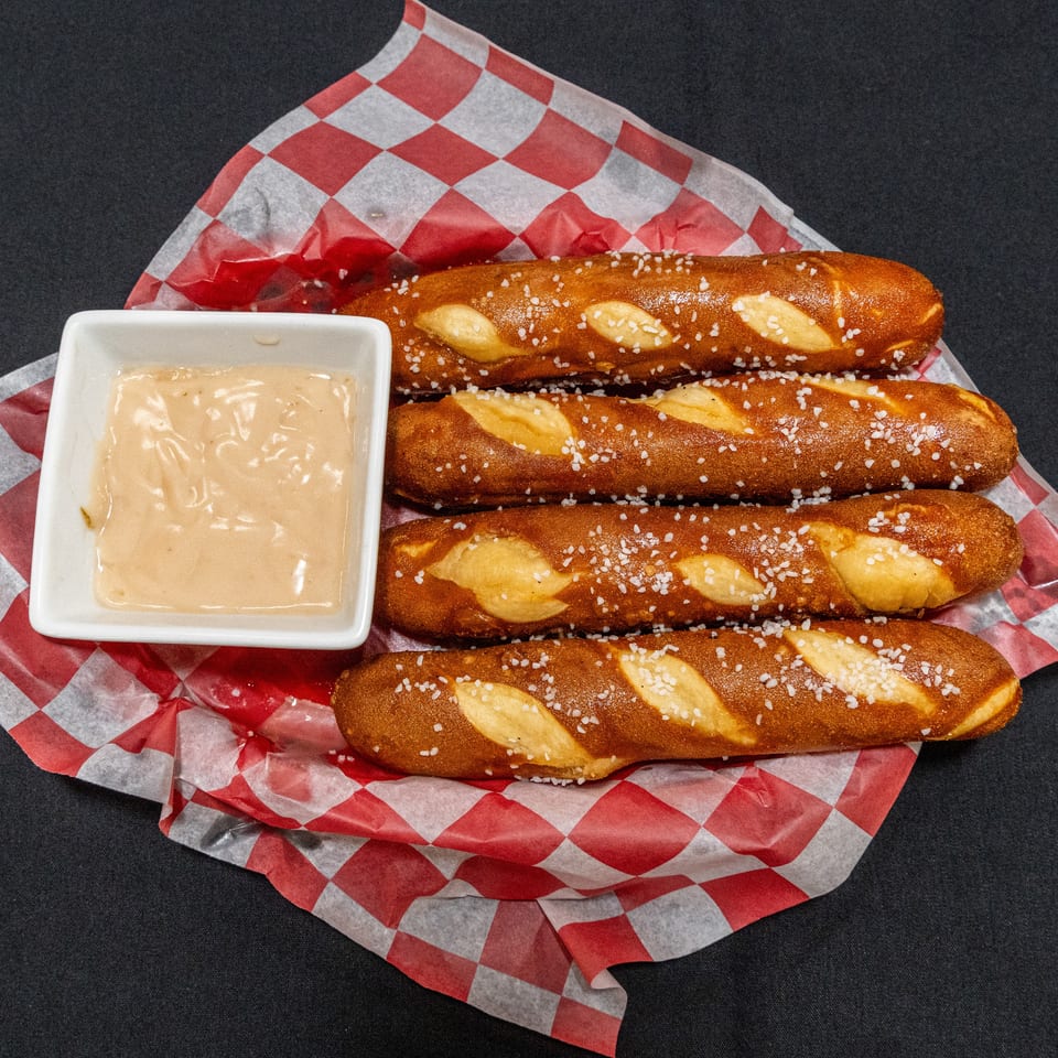 Four salted soft pretzel sticks arranged on red-and-white checkered paper with a square dish of creamy dipping sauce.