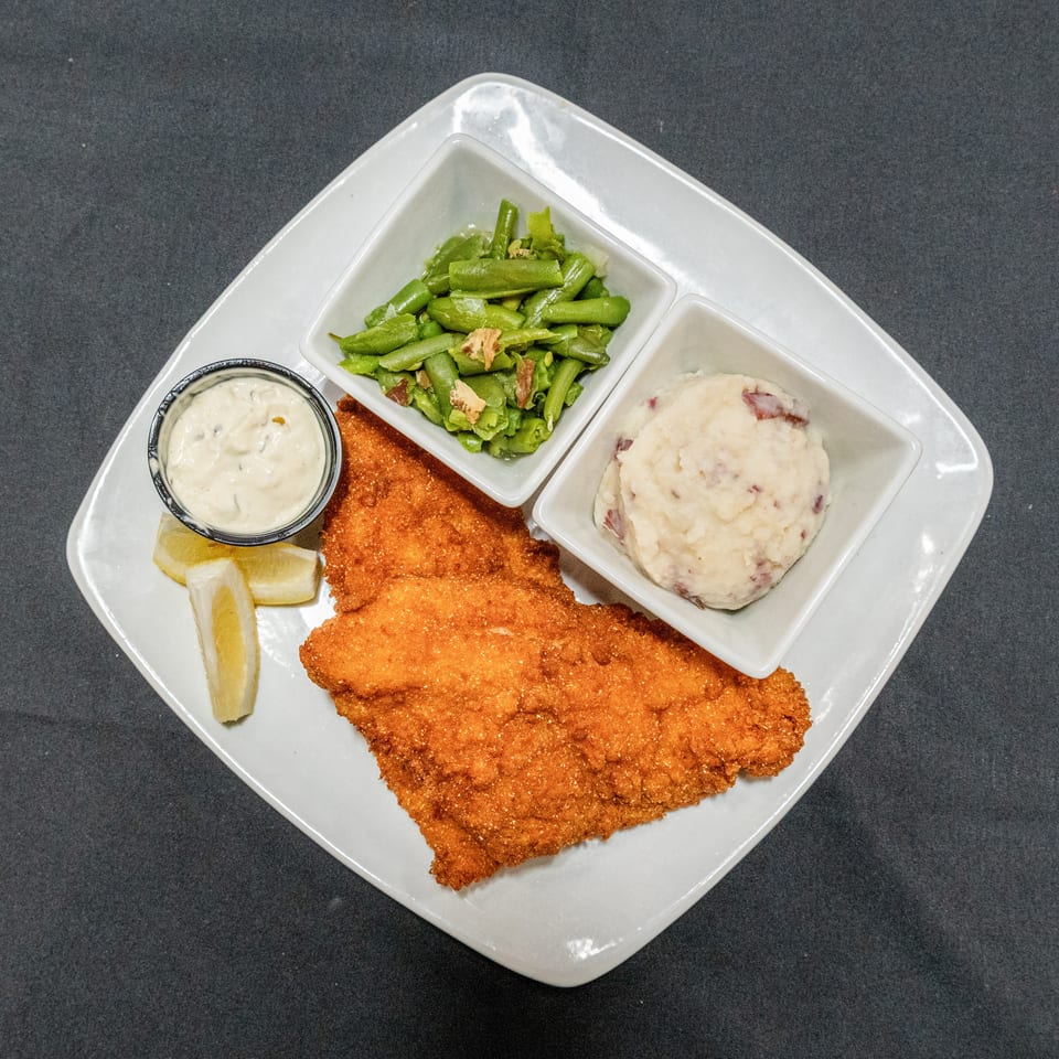 Breaded fried fish served with green beans, mashed potatoes, tartar sauce, and lemon wedges on a white plate.