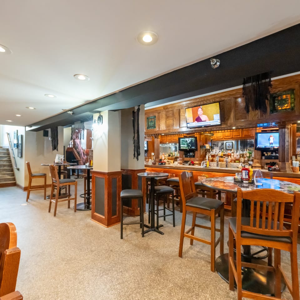 Warmly lit restaurant bar with wood paneling, televisions, liquor shelves, high-top tables, and chairs.