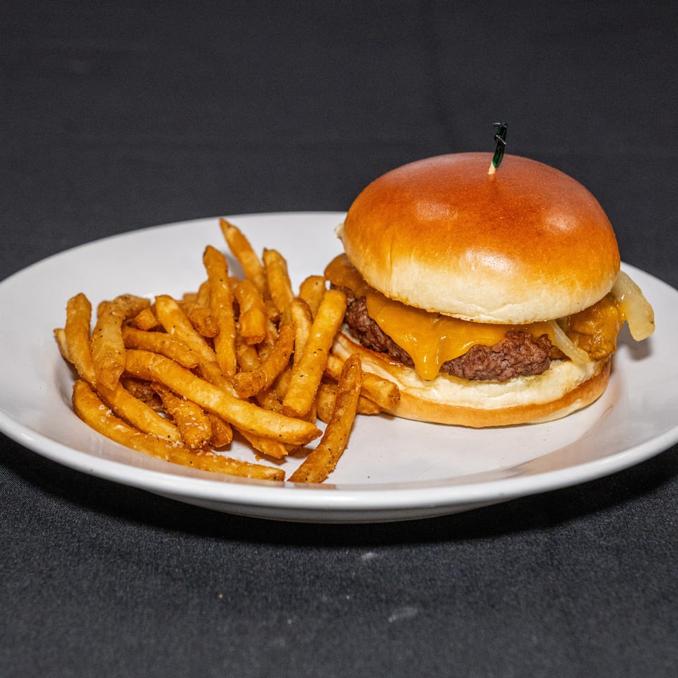 Cheeseburger with grilled onions and golden seasoned fries served on a white plate against a dark background.
